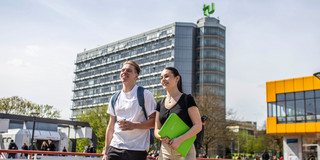 Studierende auf der Mensabrücke Ein junger Mann und eine junge Frau laufen über die Mensabrücke. Im Hintergrund sieht man den Mathetower mit TU-Logo und das Mensagebäude.