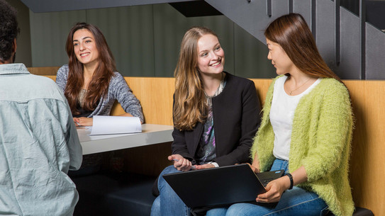 Studierende in Gesprächssituation Zwei Studierende unterhalten sich und sitzen gemeinsam auf einer Bank mit einem Laptop.