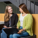 Studierende in Gesprächssituation Zwei Studierende unterhalten sich und sitzen gemeinsam auf einer Bank mit einem Laptop.