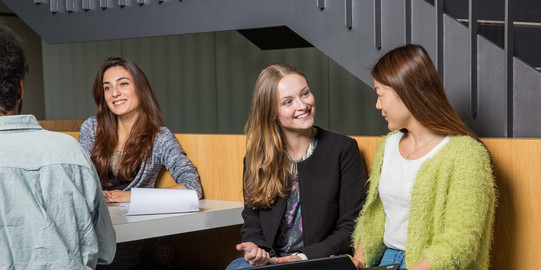 Two students are talking to each other and sitting together on a bench with a laptop.