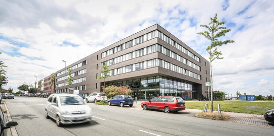 ITMC: Ein großes, braun-rot verklinkertes Gebäude mit Flachdach aus der Froschperspektive fotografiert. Davor eine Straße, auf der ein Auto fährt. Andere Autos stehen am Straßenrand hintereinander geparkt.