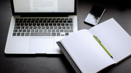Laptop, Smartphone und Notizbuch mit Stift auf einem Tisch liegend.