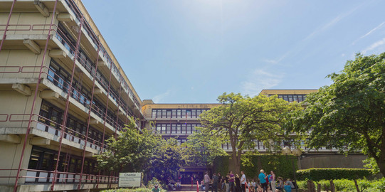 Chemiegebäude: Ein großes, breites, graues Gebäude mit Fensterfronten und Balkonen vor den Fenstern, die mit roten Metallstangen abgegrenzt werden. In der Mitte eine Treppe, auf der Menschen sitzen und links vor der Treppe Fahrräder in Fahrradständern.