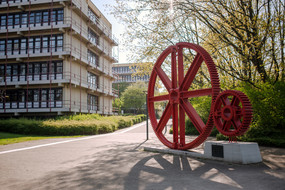 Eine rote Zahnradskulptur steht vor einem Gebäude der TU Dortmund umgeben von blühenden Bäumen im Sommer.