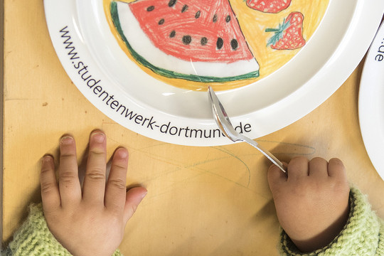 Kinderhände, die Besteck halten und auf einen Teller zeigen.