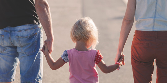 Two adults are holding each a hand of a blond child, which is seen from behind.