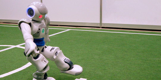 Ein Fußballroboter schießt einen roten Ball auf grünem Rasen.