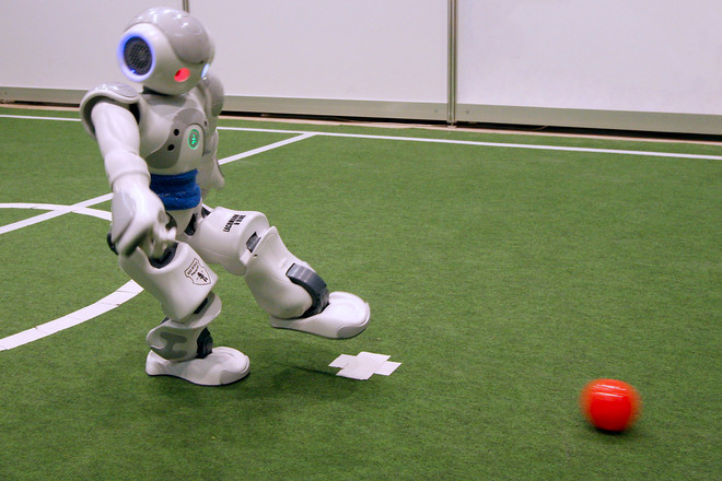 A little white robot kicking a red ball on a soccer field. 