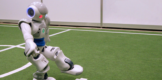 A soccer robot kicks a red ball on green grass.