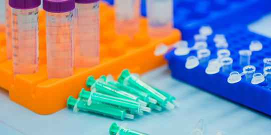 Mehrere Probenbehälter stehen in einem Fach, Zentrifugengefäße und Spritzen liegen daneben im Labor.