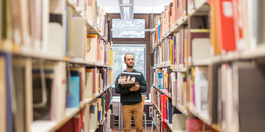 Student in der Bibliothek Studierender läuft an den Bücherregalen in der Bibliothek entlang.