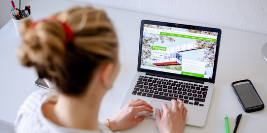 Student sitting at desk typing on laptop, books and pens next to it.