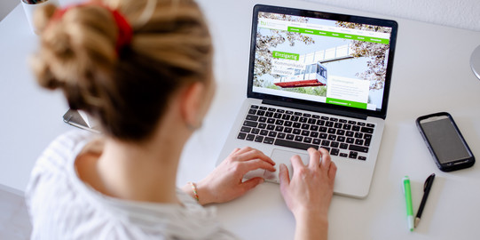 Am Schreibtisch sitzende Studentin tippt auf dem Laptop, daneben liegen Bücher und Stifte.