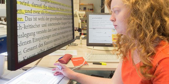 Studentin bei der Textanalyse Eine Studentin sitzt an einem Computer und liest einen Text in großer Schrift.