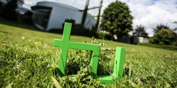 TU-Logo Im Vordergrund steht das TU-Logo im Gras, im Hintergrund das Audimax.