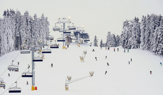 Verschneite Skipiste in Winterberg, mit Sessellift, Schneekanonen und vielen Skifahrern.
