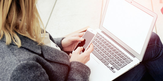 Frau mit Laptop auf dem Schoß, hält ein Smartphone in den Händen.
