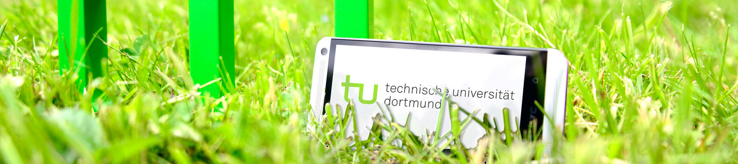Ein Smartphone, das neben dem TU-Logo im Gras steht, im Hintergrund sind die Spektralringe zu sehen.