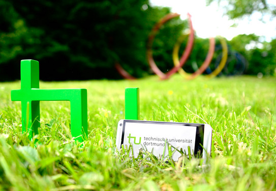TU App A smartphone standing in the grass next to the TU logo, the spectral rings can be seen in the background.