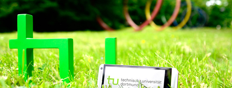 TU-App A smartphone standing in the grass next to the TU logo, the spectral rings can be seen in the background.