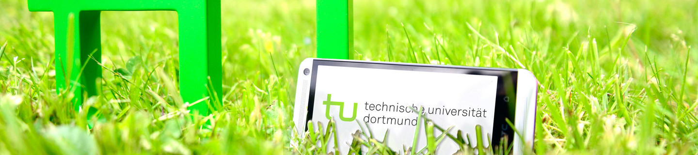 A smartphone standing in the grass next to the TU logo, the spectral rings can be seen in the background.