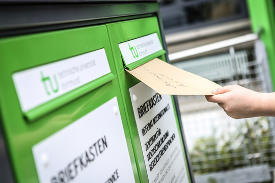 Hand of a person putting an envelope into a green mailbox with the TU Dortmund University logo on it