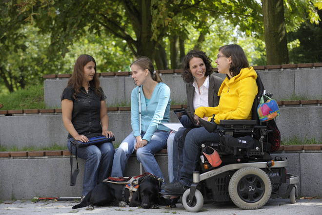 Four women sitting outside on the campus, two of them with a white cane, one of them in a wheelchair