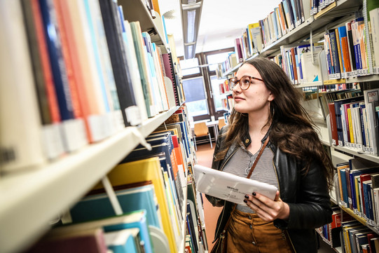 Student in the library with a tablet A student holding a tablet in her left hand is searching for a book in the library.