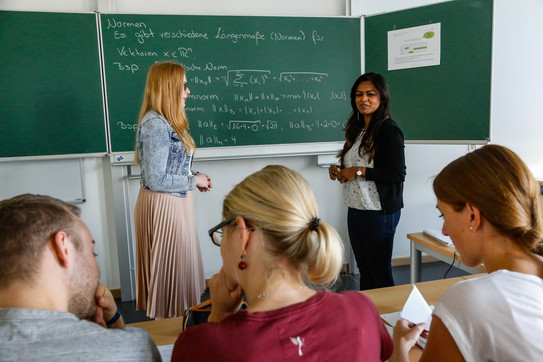 Im Hintergund stehen zwei Personen vor einer Tafel, auf die mit Kreide Formeln geschrieben sind. Im Vordegrund sieht man drei Personen von hinten, die an einem Tisch sitzen.