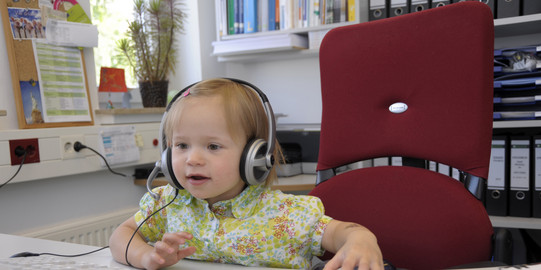 Kleinkind mit Kopfhörern bedient Maus am PC Arbeitsplatz.