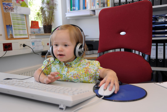 Kleinkind mit Kopfhörern bedient Maus am PC Arbeitsplatz.
