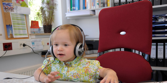 Kleinkind mit Kopfhörern bedient Maus am PC Arbeitsplatz.