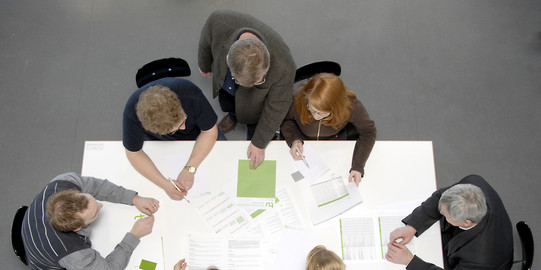 Seven people sitting around a white table with documents on it, viewed from above