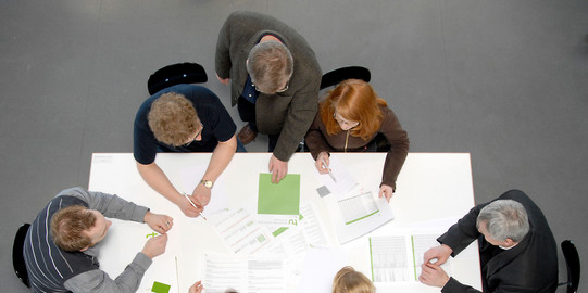 meeting situation with students and professors Seven people sitting around a white table with documents on it, viewed from above