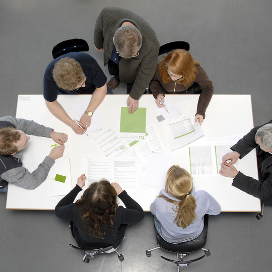 Viewed from above: Six people sitting around a table with many documents