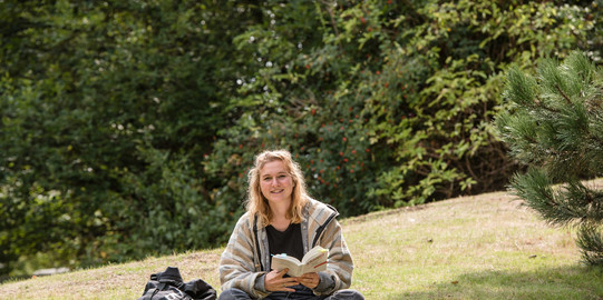 Eine Studentin sitzt im Schneidersitz auf einer Wiese mit einem Buch in der Hand.