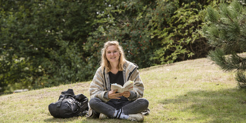 Eine Studentin sitzt im Schneidersitz auf einer Wiese mit einem Buch in der Hand.