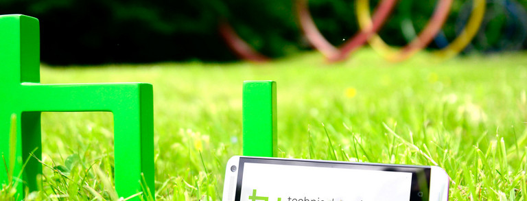 TU-App Ein grünes TU-Logo steht auf grünem Rasen. Daran lehnt ein Smartphone.