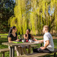 Studierende Drei Studierende sitzen an einem Sommertag an einer Sitzbank mit Tisch auf einer Wiese und unterhalten sich gut gelaunt.