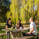Studierende Drei Studierende sitzen an einem Sommertag an einer Sitzbank mit Tisch auf einer Wiese und unterhalten sich gut gelaunt.