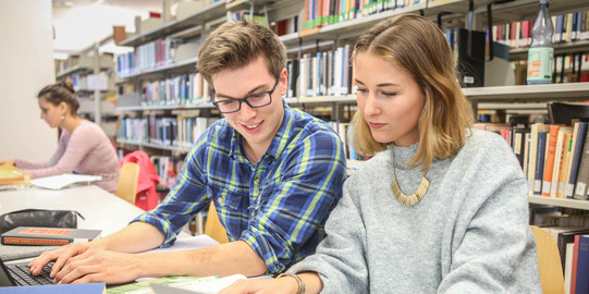 Drei Studierende sitzen an einem Tisch in der Bibliothek und arbeiten.