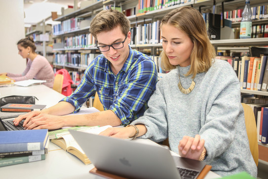 Drei Studierende sitzen an einem Tisch in der Bibliothek und arbeiten.