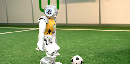 Ein Roboter mit gelbem Trikot steht mit einem Ball am Anstoßpunkt.