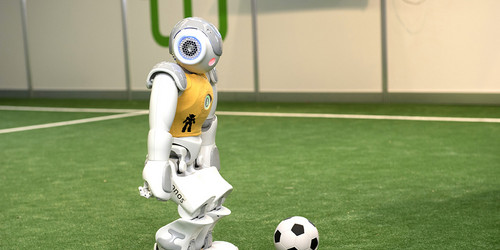 Ein Roboter mit gelbem Trikot steht mit einem Ball am Anstoßpunkt.