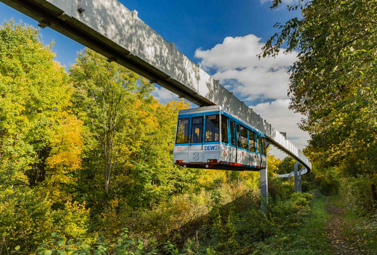 The H-Bahn runs along a path with green trees.