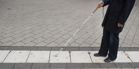 Eine blinde Person folgt dem Blindenleitsystem auf dem Campus mithilfe eines Blindenstocks