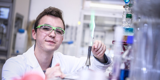 Männlicher Auszubildender mit Kittel und Schutzbrille im Chemielabor erzeugt eine grüne Flamme am Bunsenbrenner.