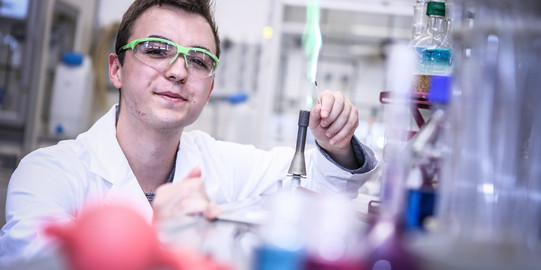 Männlicher Auszubildender mit Kittel und Schutzbrille im Chemielabor erzeugt eine grüne Flamme am Bunsenbrenner.