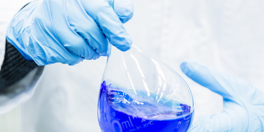 Zwei Hände in blauen Gummihandschuhen halten einen Messkolben, der mit blauer Flüssigkeit gefüllt ist.