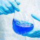 Zwei Hände in blauen Gummihandschuhen halten einen Messkolben, der mit blauer Flüssigkeit gefüllt ist.